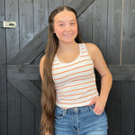 Young girl with long hair wearing a striped tank top and jeans standing in front of a wooden door.