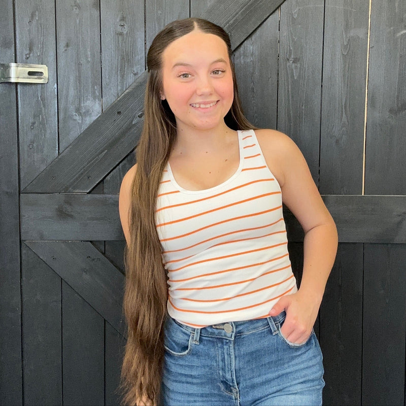 Young girl with long hair wearing a striped tank top and jeans standing in front of a wooden door.