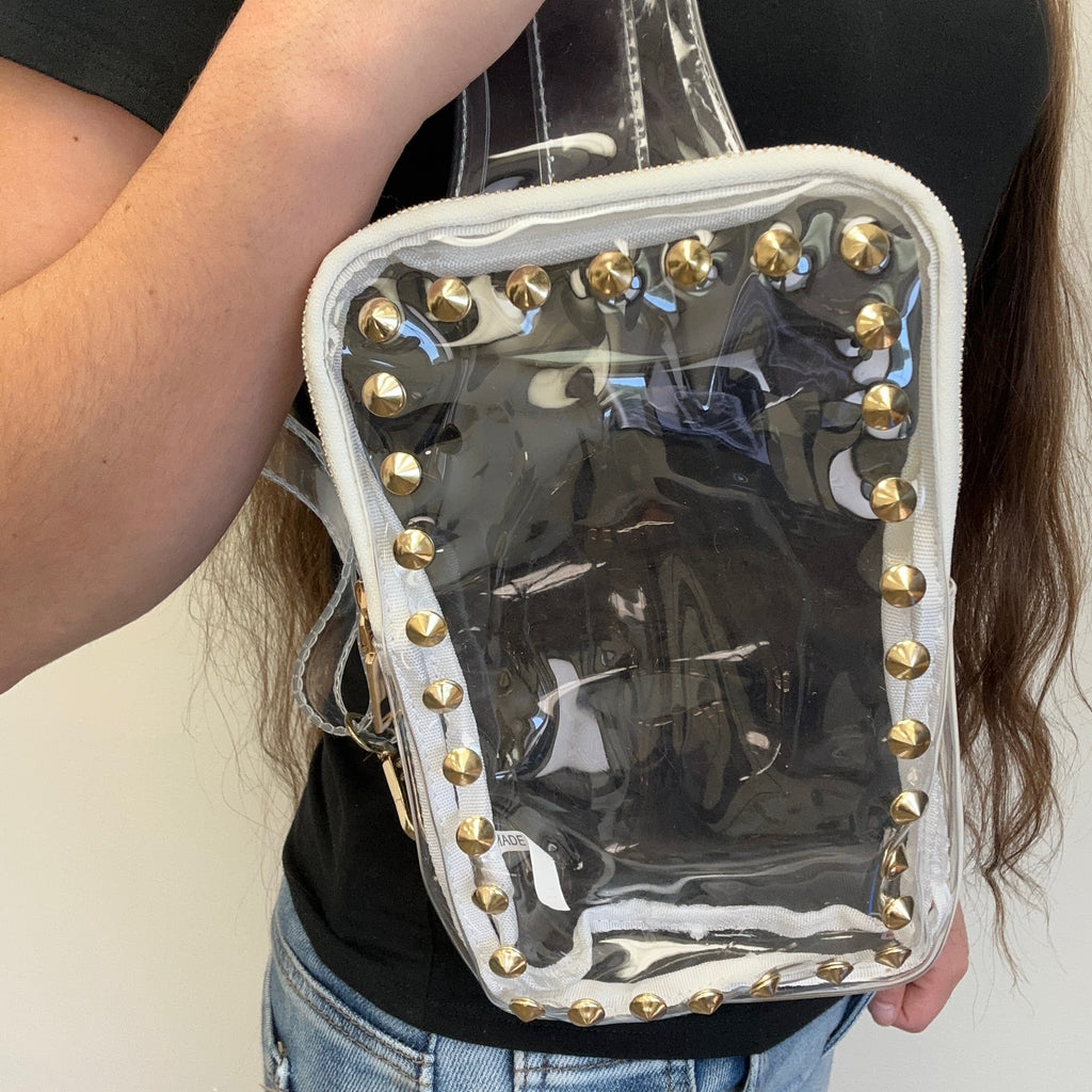Clear handbag with gold studs held by a person against a neutral background