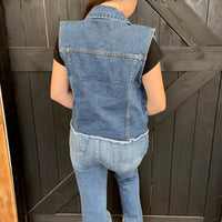 Person wearing a denim vest and jeans standing against a wooden background