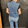 Person wearing a denim vest and jeans standing against a wooden background