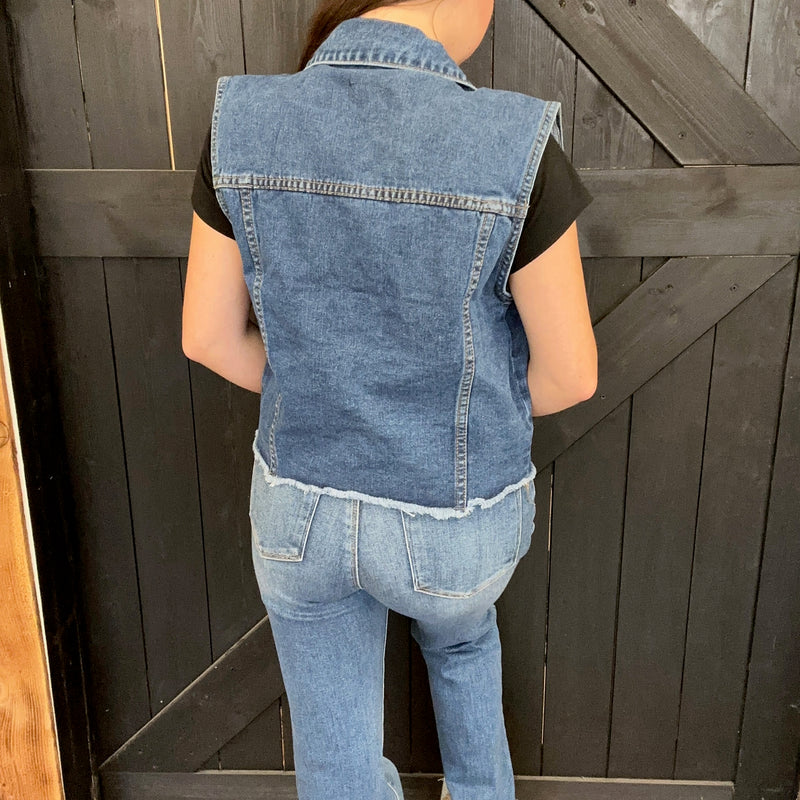 Person wearing a denim vest and jeans standing against a wooden background