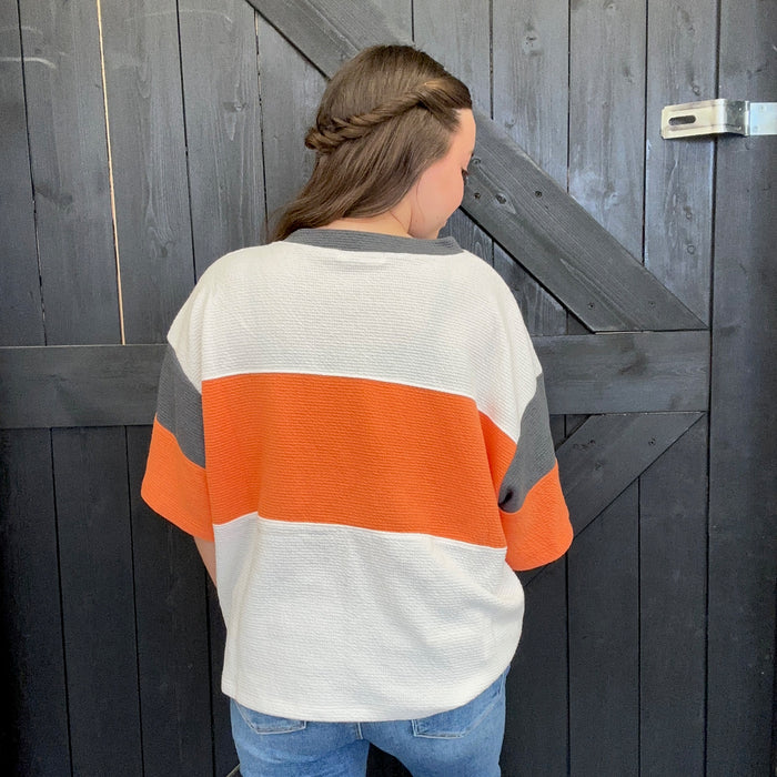 Striped sweater with orange and gray colors, standing against a wooden door.