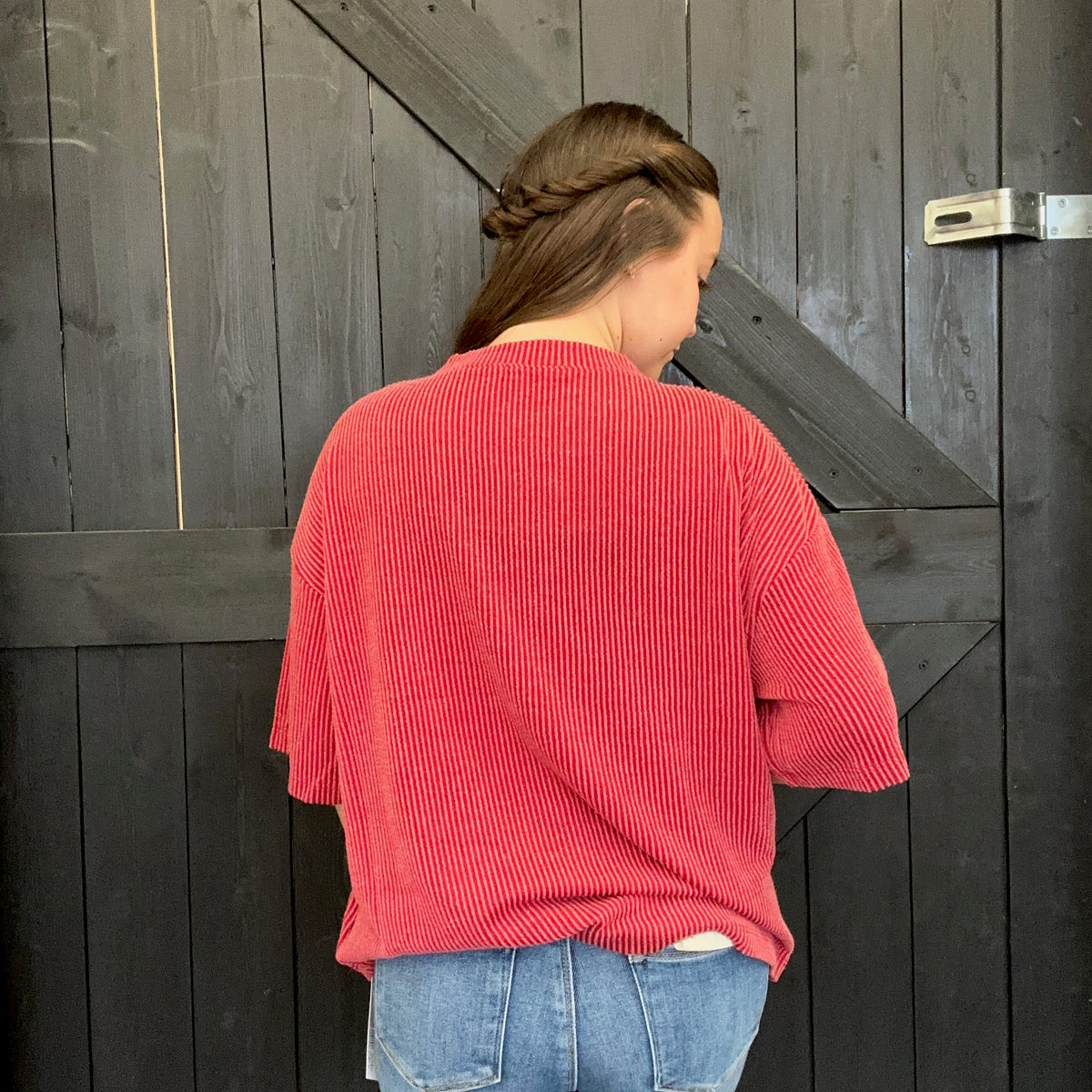 Person wearing a red ribbed sweater and blue jeans standing against a wooden wall.