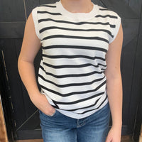 Person wearing a black and white striped sleeveless top and blue jeans against a dark tiled wall.