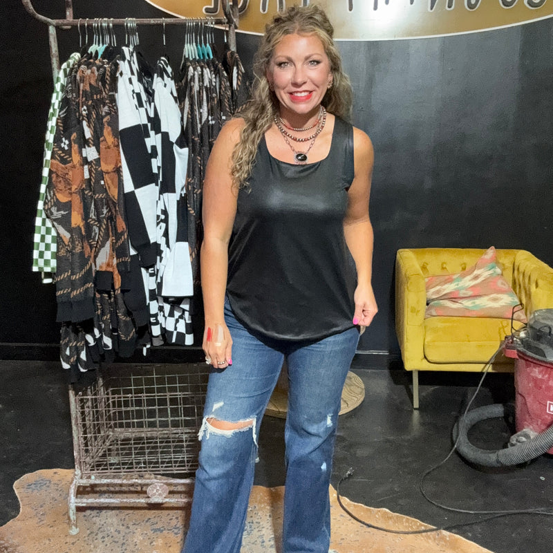 Woman in black tank top and jeans standing in front of a rack of clothes with a yellow couch and sign in the background.