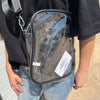 Clear crossbody bag with a visible brand label held by a person wearing a black shirt and blue jeans.