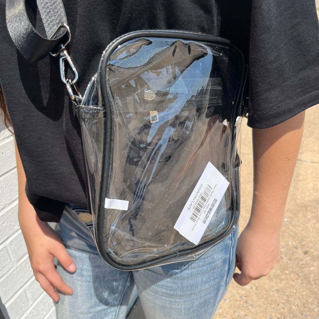 Clear crossbody bag with a visible brand label held by a person wearing a black shirt and blue jeans.
