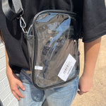 Clear crossbody bag with a visible brand label held by a person wearing a black shirt and blue jeans.