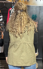 Person wearing a green vest in front of a rack of clothes with a yellow chair and blackboard in the background.