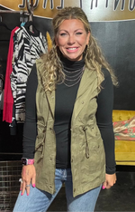 Woman wearing a green vest and black top in a store setting with clothing racks and a yellow couch.