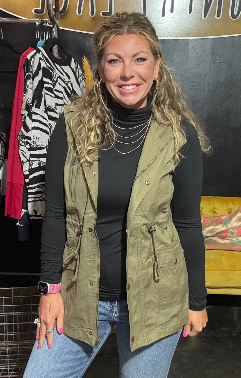 Woman wearing a green vest and black top in a store setting with clothing racks and a yellow couch.