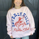 Person wearing a pink 'Beverly Hills Rodeo Club' sweatshirt against a wooden background