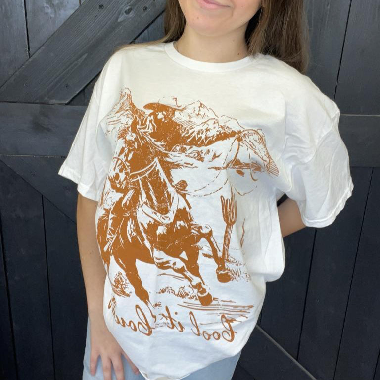 Person wearing a white t-shirt with a graphic design of a person on a horse and text, standing against a dark wooden background.