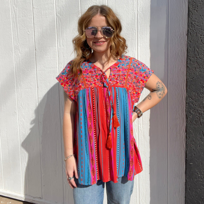 A woman standing in front of a white wall wearing a colorful embroidered top with short sleeves and a round neckline.