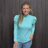 Woman wearing a light blue turtleneck sweater standing in a clothing store with racks of clothes and a yellow chair in the background.
